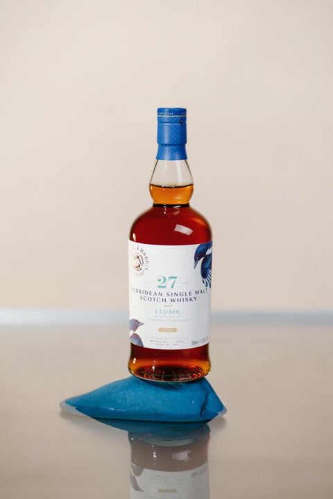 A bottle of Ferg and Harris Ledaig, placed on top of a blue rock surrounded by still water, with bottle reflection on water