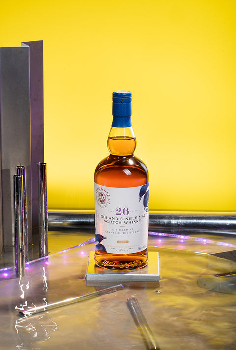 A bottle of Ferg and Harris 26-year-old, placed on a tile on top of water. In front of a bright yellow background with metallic details and bright purple light strip