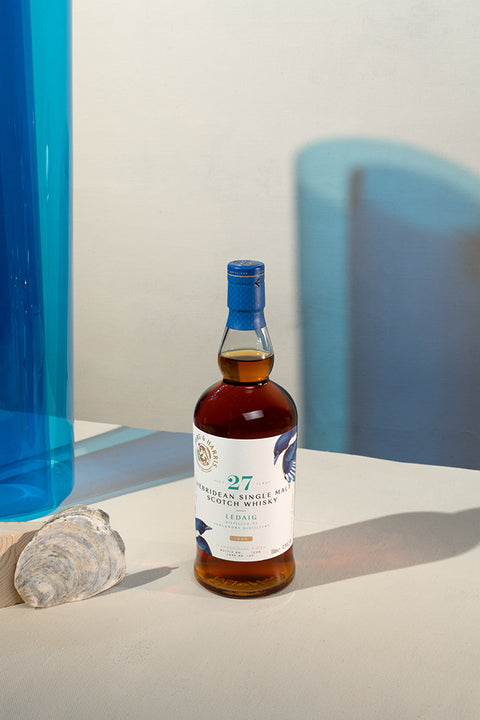 A bottle of Ferg and Harris 27-year-old Ledaig, with seashells next to it and blue transparency and shadow in the background.